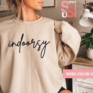 May include: A beige sweatshirt with the text "indoorsy" written in black cursive script. The sweatshirt is being worn by a person. The text "MORE COLOR OPTIONS" is visible in the bottom right corner of the image.