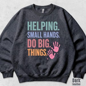 May include: A dark heather gray crewneck sweatshirt featuring the text HELPING. SMALL HANDS. DO BIG. THINGS. in light blue, lavender, pink, and orange, accompanied by two pink handprint graphics on the front center of the garment.
