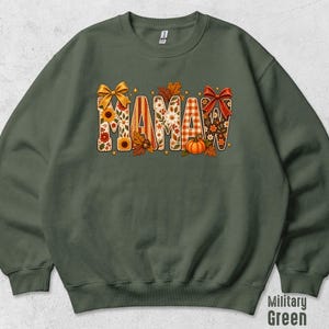 May include: A military green crewneck sweatshirt featuring the word MAMA in large block letters. The letters are decorated with autumn-themed patterns including sunflowers, orange plaid, fall leaves, acorns, a pumpkin, and two red bows.