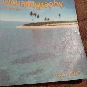 Può includere: Un libro intitolato "Invitation to Oceanography, Fifth Edition" di Paul R. Pinet. La copertina presenta un'isola tropicale con palme e un oceano blu.