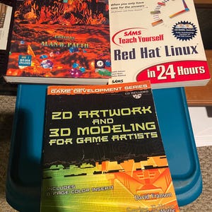 May include: Three technical books titled Graphics Gems V edited by Alan W. Paeth, Sams Teach Yourself Red Hat Linux in 24 Hours, and 2D Artwork and 3D Modeling for Game Artists by David Franson, featuring colorful graphic covers and text.
