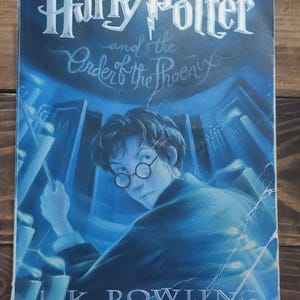 May include: A book cover for "Harry Potter and the Order of the Phoenix" by J.K. Rowling. The cover features a blue-toned illustration of Harry Potter with glasses, looking over his shoulder. The title is in a stylized font.