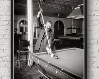 May include: A black and white photograph of a man playing snooker. He is standing at a snooker table and lining up a shot with a cue stick. There are two snooker balls on the table, one white and one red.