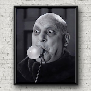 May include: A black and white framed print featuring a bald individual with dark eyes and a prominent brow, holding a glowing light bulb in their mouth. The subject wears a dark, textured garment, and the image is presented in a simple black frame.