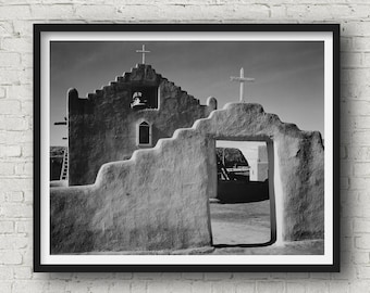 May include: Black and white photograph of a church building with a cross on top. The church is made of adobe brick and has a large arched doorway. The photo is framed in a black frame.