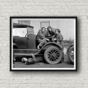 May include: Black and white framed print of four women working on a vintage car. Three women are standing around the car, while one is working underneath. The car has a spare tire and wooden spoke wheels. The women are wearing coveralls.