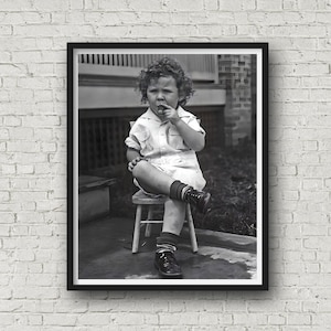 May include: A black and white photo of a young child sitting on a small wooden stool. The child is wearing a white shirt, shorts, and socks. They are holding a small object in their hand and looking at it.