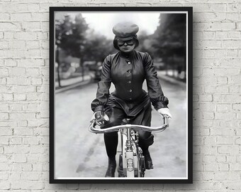 Könnte beinhalten: Schwarz-Weiß-Druck einer Frau auf einem Vintage-Fahrrad. Sie trägt ein dunkles, geknöpftes Kleid, eine Mütze und eine Schutzbrille. Das Fahrrad hat einen geschwungenen Lenker und einen Frontscheinwerfer. Der Hintergrund zeigt eine von Bäumen gesäumte Straße.