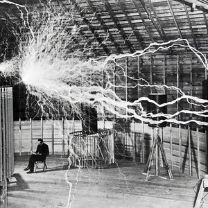 May include: Black and white photograph of a man seated in a chair, observing a large electrical experiment. A spherical object emits a massive discharge of electricity, with lightning-like arcs extending across the room. The scene is set in a wooden structure.