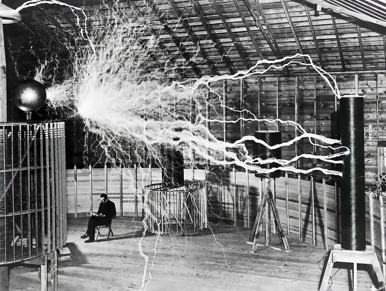 May include: Black and white photograph of a man seated in a chair, observing a large electrical experiment. A spherical object emits a massive discharge of electricity, with lightning-like arcs extending across the room. The scene is set in a wooden structure.