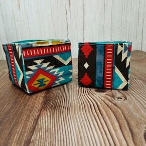 May include: Two small, square fabric storage containers with a Southwestern geometric pattern in red, turquoise, black, and cream. The containers are open at the top and sit on a wooden surface.
