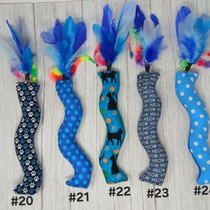 May include: Five fabric cat toys shaped like wavy tubes, each topped with blue and purple feathers and a colorful spring. Patterns include paw prints, blue polka dots, black cat silhouettes, the word MEOW, and light blue polka dots, labeled 20-24.