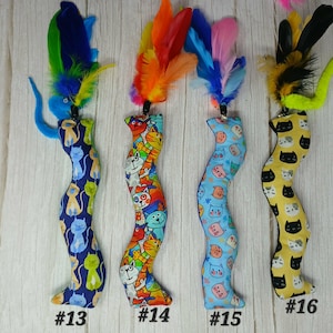 May include: Four cat toys with colorful feather accents. Each toy has a unique patterned fabric body, including blue with cat silhouettes, a vibrant cat-face print, light blue with cat faces, and yellow with black cat faces. Each toy is labeled with a number.
