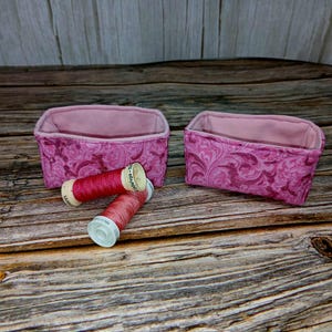 May include: Two small, rectangular fabric baskets with a pink floral pattern, and two spools of pink thread. The baskets are lined with a solid pink fabric. The spools of thread are red and pink.