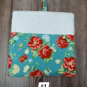 May include: A teal blue pot holder with a floral pattern featuring red roses and white accents. The pot holder has a white lining and a loop for hanging.