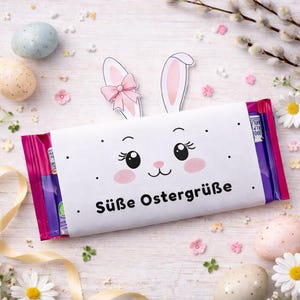 Könnte beinhalten: Eine weiße, österliche Schokoriegelverpackung mit einem Hasengesicht, rosa Wangen und Wimpern sowie dem Text "Süße Ostergrüße". Umgeben von Ostereiern, Blumen und einem gelben Band.