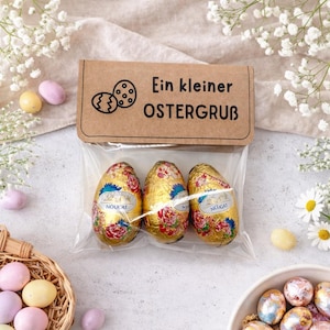 Può includere: Tre uova di Pasqua di cioccolato avvolte in carta dorata con disegni floreali, confezionate in un sacchetto trasparente. La confezione include un biglietto marrone con il testo "Ein kleiner OSTERGRUB" e illustrazioni di uova di Pasqua.