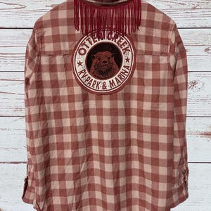 This flannel shirt has a patch like graphic on the back that says " Otter Creek RV Park & Marina, and some fringe under the collar.