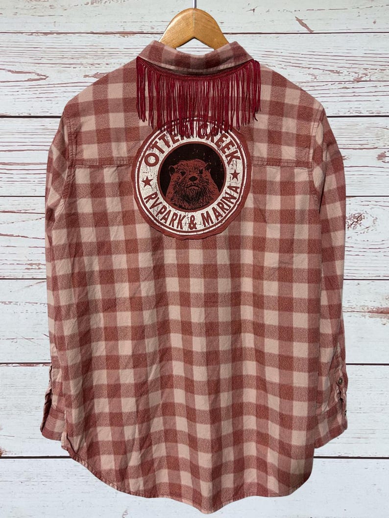 This flannel shirt has a patch like graphic on the back that says " Otter Creek RV Park & Marina, and some fringe under the collar.