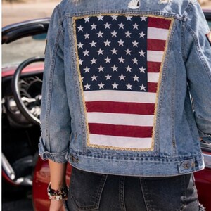 May include: Light blue denim jacket with a large American flag design on the back. The flag features white stars on a blue background and red and white stripes. The jacket has a distressed look.