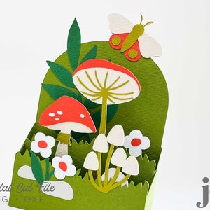 May include: A green cardstock box with a cut-out design of mushrooms, leaves, and a butterfly. The mushrooms are red and white, and the butterfly is white with red and green accents. The text "Digital Cut File SVG  DXF" is printed on the bottom of the box.