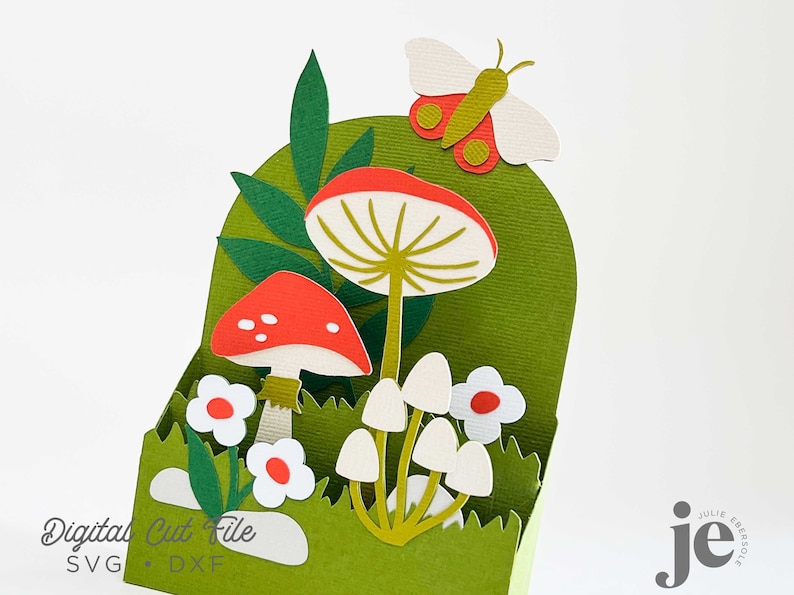 May include: A green cardstock box with a cut-out design of mushrooms, leaves, and a butterfly. The mushrooms are red and white, and the butterfly is white with red and green accents. The text "Digital Cut File SVG  DXF" is printed on the bottom of the box.