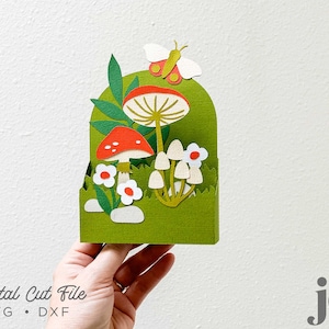 May include: A green papercraft box with a mushroom and butterfly design. The box is decorated with red and white mushrooms, green leaves, and a white butterfly. The box is a digital cut file, available in SVG and DXF formats.