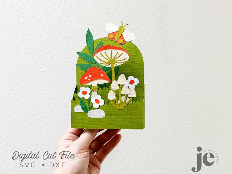 May include: A green papercraft box with a mushroom and butterfly design. The box is decorated with red and white mushrooms, green leaves, and a white butterfly. The box is a digital cut file, available in SVG and DXF formats.