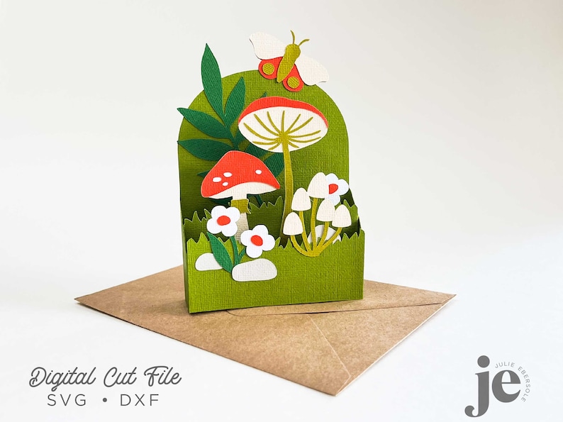 May include: A green papercraft card with a pop-up scene of mushrooms, flowers, and a butterfly. The card is on a brown envelope. The card is a digital cut file, available in SVG and DXF formats.