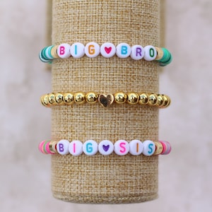 May include: Three bracelets with white beads and colorful accents. The top bracelet spells "BIG BRO" with a red heart bead. The middle bracelet is gold. The bottom bracelet spells "BIG SIS" with a purple heart bead.