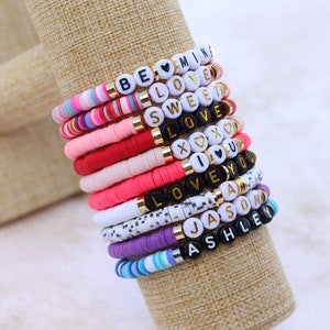 May include: A stack of colourful beaded bracelets with different words spelled out on them, including "Be Mine", "Love", "Sweet", "Love", "XOXO", "I Love You", "Love", "Jason", and "Ashley".