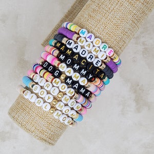 Pode incluir: Uma pilha de pulseiras de contas coloridas com diferentes palavras escritas em letras douradas. As palavras incluem "Mom", "Girl", "Boy", "Mama", "Mommy", "Dog", "Amy", "Madre", "Mami" e "Mam&aacute;cita".