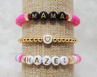 May include: Three beaded bracelets. The top bracelet has black letter beads spelling "MAMA" on a pink base. The middle bracelet is gold with a white heart bead. The bottom bracelet spells "HAZEL" with white letter beads on a colorful base.