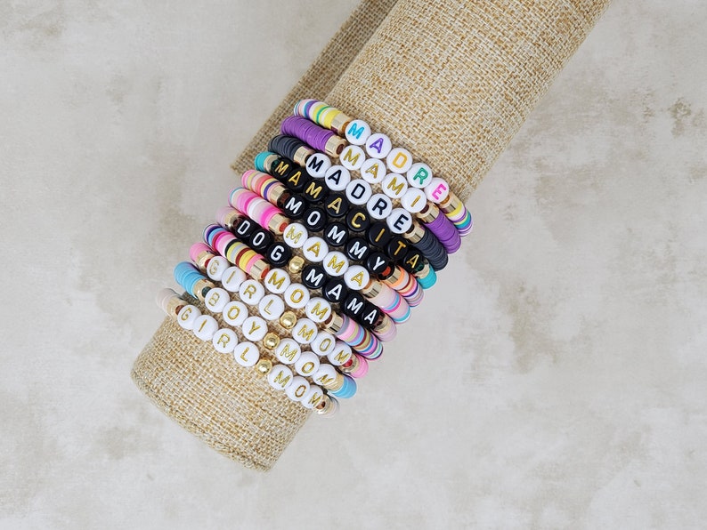 Pode incluir: Uma pilha de pulseiras de contas coloridas com diferentes palavras escritas em letras douradas. As palavras incluem "Mom", "Girl", "Boy", "Mama", "Mommy", "Dog", "Amy", "Madre", "Mami" e "Mam&aacute;cita".