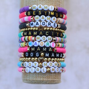Pode incluir: Uma pilha de pulseiras de contas coloridas com diferentes palavras escritas nelas, incluindo "Mami", "Best Mom", "Mama", "Madre", "Mamacita", "Mom", "Dog Mama", "Boy Mom" e "Girl Mom".
