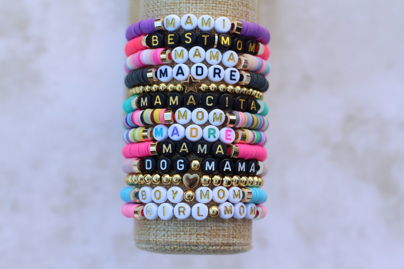 Pode incluir: Uma pilha de pulseiras de contas coloridas com diferentes palavras escritas nelas, incluindo "Mami", "Best Mom", "Mama", "Madre", "Mamacita", "Mom", "Dog Mama", "Boy Mom" e "Girl Mom".