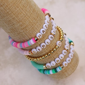 May include: Stack of colorful beaded bracelets with white letter beads spelling out "HAZE", "GIRLS", and "SISTER". The bracelets are displayed on a tan fabric surface.