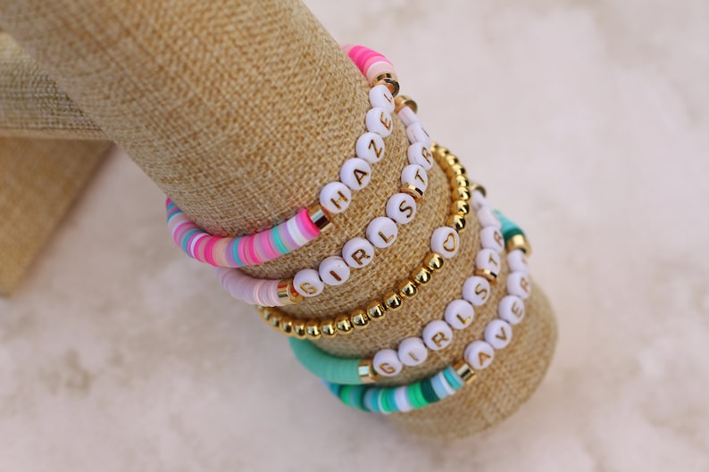 May include: Stack of colorful beaded bracelets with white letter beads spelling out "HAZE", "GIRLS", and "SISTER". The bracelets are displayed on a tan fabric surface.