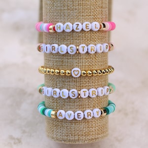 May include: Five colorful beaded bracelets with white letter beads spelling out names and phrases. The bracelets include gold accents and are displayed on a neutral-toned cylindrical stand. The names "HAZEL" and "AVERY" are visible.