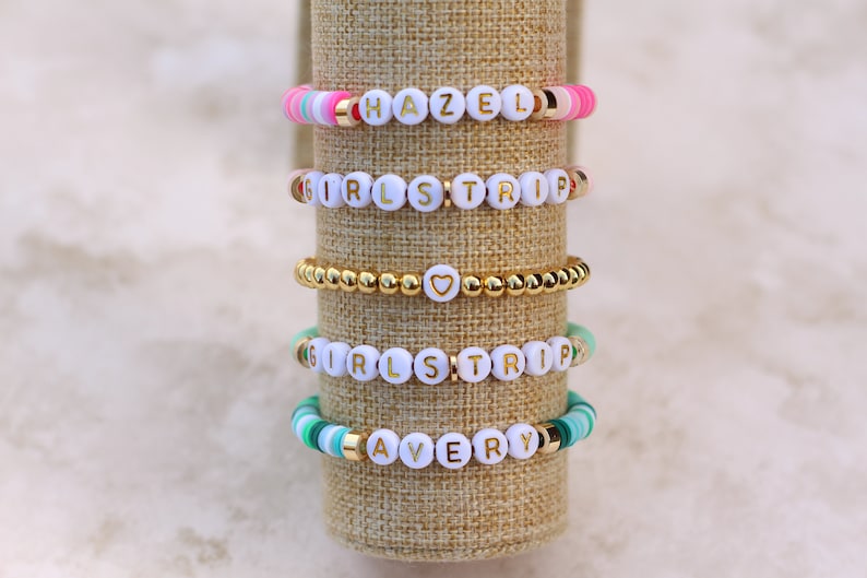 May include: Five colorful beaded bracelets with white letter beads spelling out names and phrases. The bracelets include gold accents and are displayed on a neutral-toned cylindrical stand. The names "HAZEL" and "AVERY" are visible.