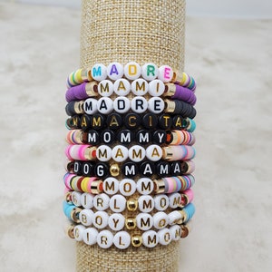Pode incluir: Uma pilha de pulseiras de contas coloridas com diferentes palavras escritas nelas, incluindo "MADRE", "MAMI", "MAMACITA", "MOMMY", "MAMA", "DOG MAMA", "MOM", "GIRL MOM", "BOY MOM" e "GIRL MOM".