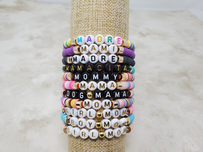 Pode incluir: Uma pilha de pulseiras de contas coloridas com diferentes palavras escritas nelas, incluindo "MADRE", "MAMI", "MAMACITA", "MOMMY", "MAMA", "DOG MAMA", "MOM", "GIRL MOM", "BOY MOM" e "GIRL MOM".