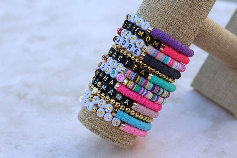 Pode incluir: Uma pilha de pulseiras de contas coloridas com diferentes palavras escritas nelas, incluindo "Mami", "Mom", "Grandma" e "Girl".