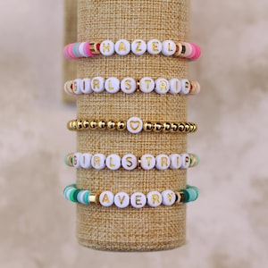 May include: Five bracelets stacked on a brown fabric display stand. The bracelets are made of white beads with gold lettering. The top bracelet spells "HAZEL", the second bracelet spells "GIRLSTRIP", the third bracelet is gold beads with a gold heart charm, the fourth bracelet spells "GIRLSTRIP", and the bottom bracelet spells "AVERY".