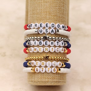 May include: A stack of colorful beaded bracelets with patriotic themes. The bracelets feature white beads with black lettering spelling out "FREEDOM", "USA", "LIBERTY", "AMERICA", "MERICA", and "1776".