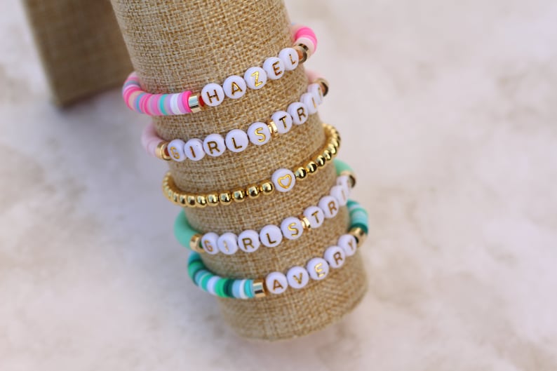 May include: Stack of colorful beaded bracelets with white letter beads spelling out names and phrases. The bracelets feature gold accents and a variety of colors, including pink, blue, and green. The bracelets are displayed on a tan fabric.