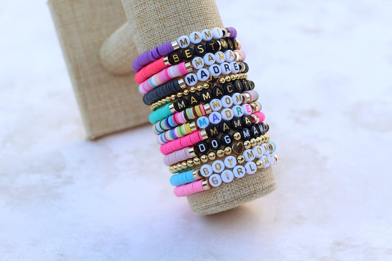 Pode incluir: Uma pilha de pulseiras de contas coloridas com diferentes palavras escritas nas contas, incluindo "Mami", "Best", "Mama", "Madre", "Mom", "Girl", "Boy" e "Dog".