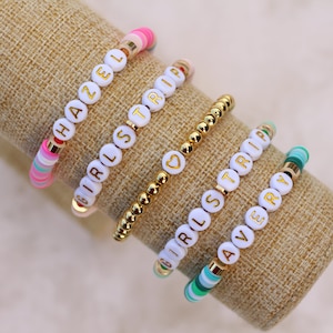 May include: A collection of beaded bracelets with white letter beads spelling out names and phrases. The bracelets feature gold accents and colorful beads in pink, green, and blue. The bracelets are displayed on a tan fabric.