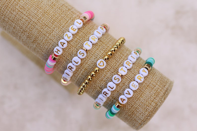 May include: A collection of beaded bracelets with white letter beads spelling out names and phrases. The bracelets feature gold accents and colorful beads in pink, green, and blue. The bracelets are displayed on a tan fabric.