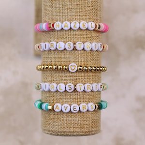May include: Five beaded bracelets with white letter beads spelling "HAZEL", "GIRLS TRIP", and "AVERY". The bracelets feature gold and colorful beads in pink, green, and gold. One bracelet is all gold beads with a heart charm.